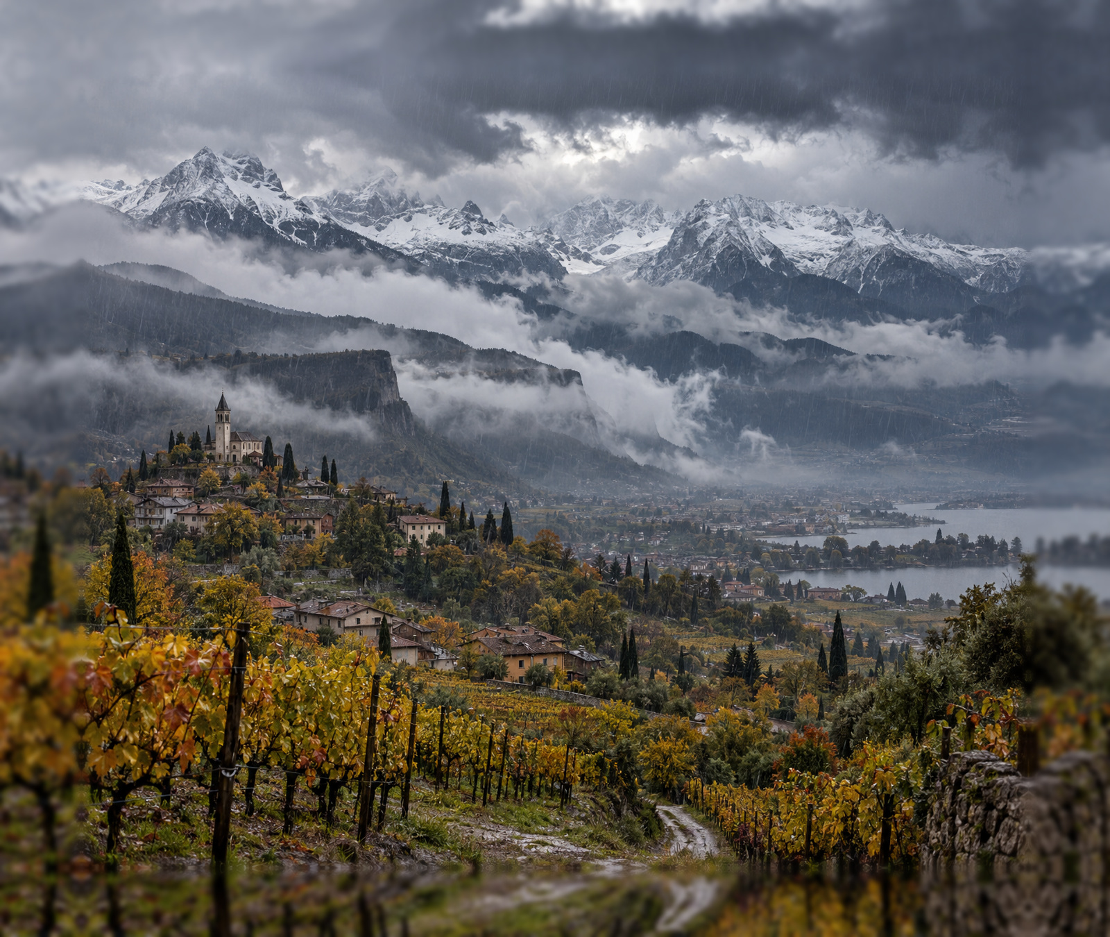 Paesaggio alpino italiano in autunno con nubi basse, pioggia e neve sulle cime più alte.