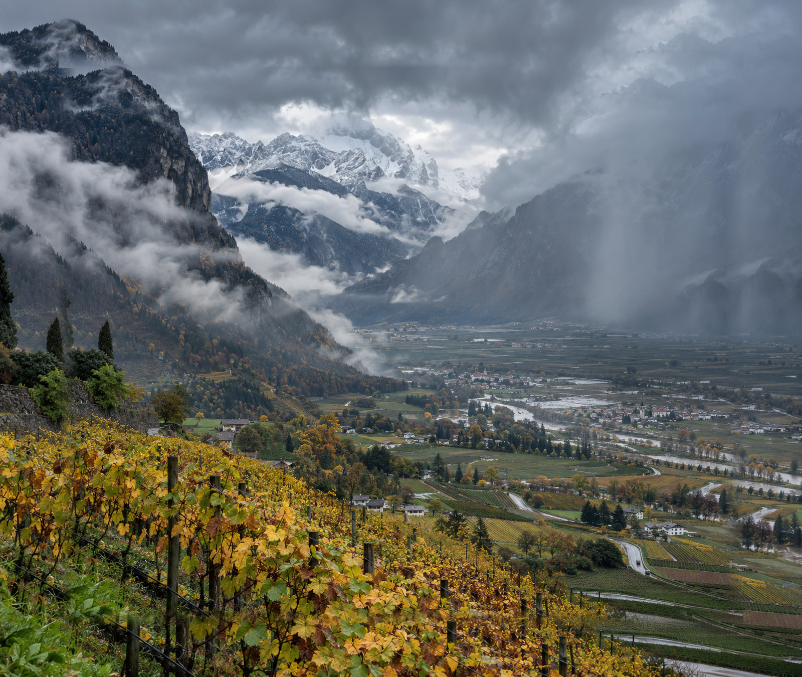 Valle alpina italiana in autunno sotto nubi basse e pioggia, con neve fresca solo sulle cime più alte.